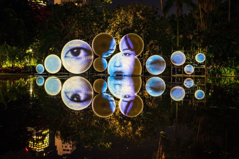 Projected eyes and faces arranged across circular screens in a garden at night.